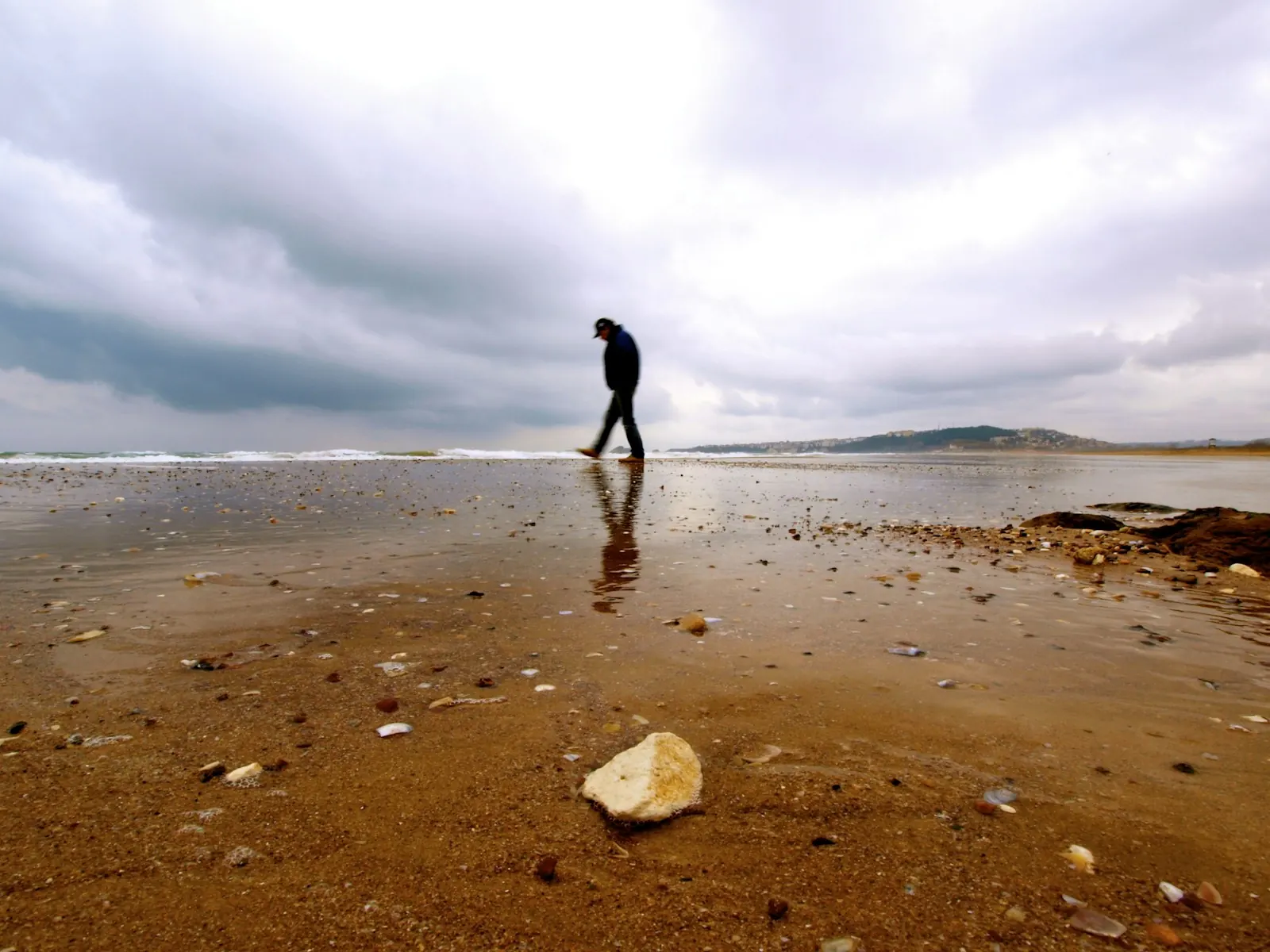 Person walking alone on a wet sandy beach under a cloudy sky