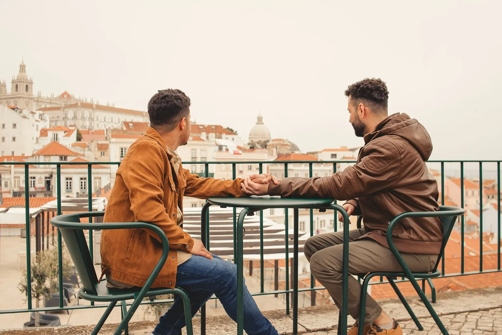 Two men holding hands at a cafe table overlooking city rooftops