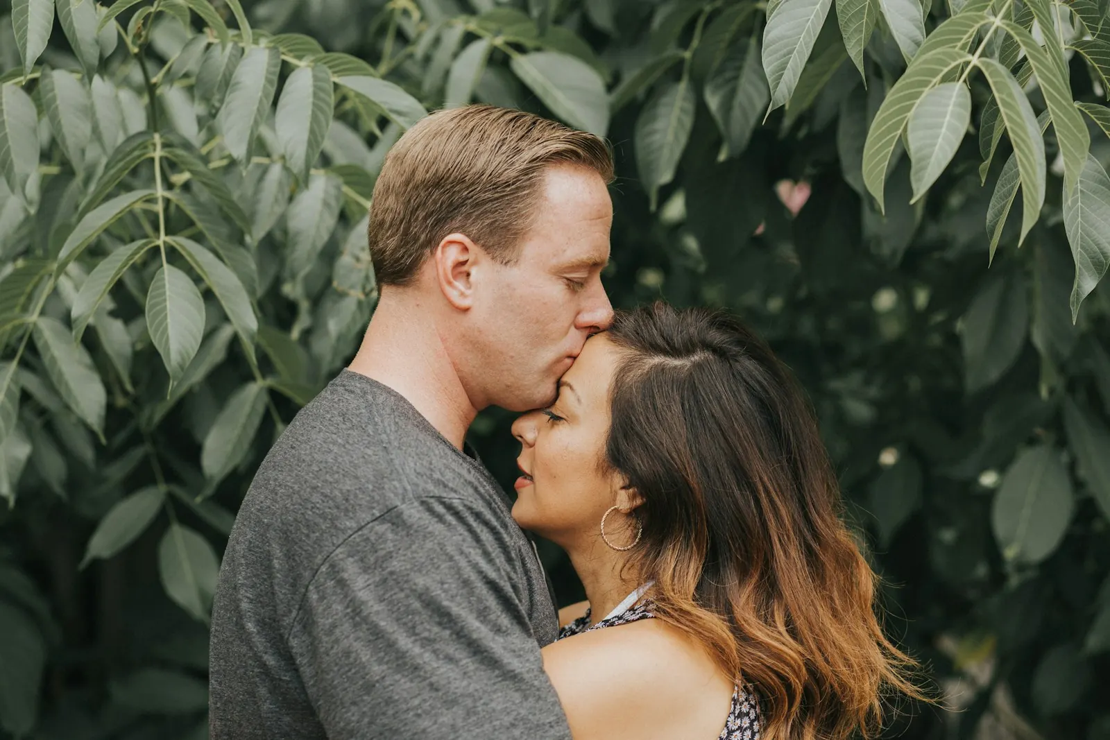 Man tenderly kissing his partner's forehead among green leaves